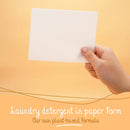 Eine Hand hält ein weißes Waschmittelblatt vor beigem Hintergrund, betont "Waschmittel in Papierform" und "Unsere eigene pflanzliche Formel".