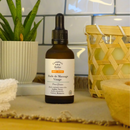 Eine bernsteinfarbene Tropfflasche Gesichtsmassageöl für fettige Haut auf einer Theke mit Pflanzen und Handtüchern.