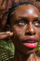  Nahaufnahme des Gesichts einer Frau mit leuchtenden Augen, die eine erdige Gesichtsmaske auf ihre Wange aufträgt, vor einem Hintergrund aus grünen Blättern.