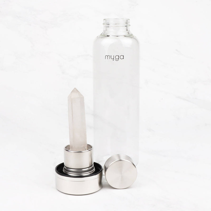 Eine klare Glaswasserflasche mit herausgenommenem Kristall-Infuser und daneben liegendem Deckel auf einer weißen Marmorfläche, die das „myga“-Logo zeigt.