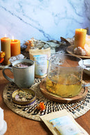 Eine Glasteekanne mit Kräutertee, eine Tasse und eine kleine Schale mit getrockneten Kräutern stehen auf einem Tisch, umgeben von Kerzen und getrockneten Blumen.