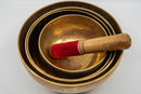 Ein Satz von vier gestapelten Klangschalen aus Messing, von oben gesehen, mit einem Holzschlägel mit roter Samtspitze in der kleinsten Schale.
