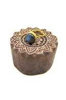 Ein goldener Fingerring mit einem runden, blaugrauen Labradorit-Edelstein auf einem dunklen, handgeschnitzten Holzblock mit floralem Mandala-Muster.
