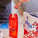 Nahaufnahme einer Person, die Pulver aus einem kleinen roten ARGI+-Beutel in eine ebenfalls mit ARGI+ gebrandete rote Sportflasche füllt. Eine größere Tüte des Nahrungsergänzungsmittels L-Arginin- & Vitaminkomplex ist im Hintergrund sichtbar.