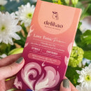 Hände halten eine Delikao Love Tonic Passion Zartbitterschokolade vor einem Hintergrund aus weißen Blumen.