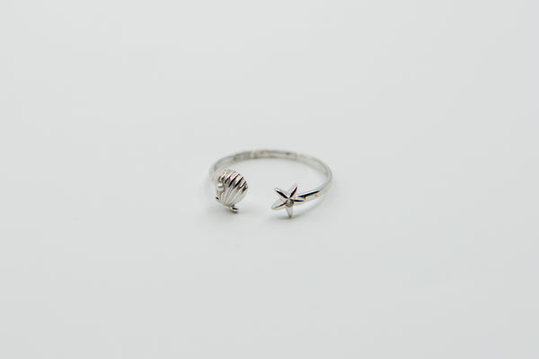 Ein offener silberner Ring mit einem Muschel-Anhänger an einem Ende und einem Stern-Anhänger mit einem kleinen Stein am anderen, auf einem weißen Hintergrund.