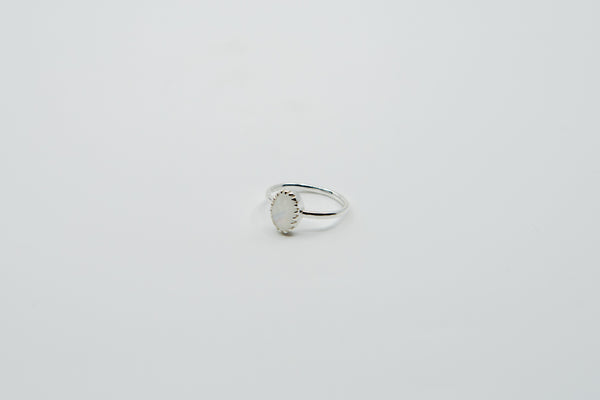 Ein silberner Ring mit einer schlichten Ringschiene und einem ovalen, irisierenden weißen Stein, der von einem zarten gewellten Rand umgeben ist, auf einem weißen Hintergrund.