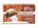 Eine Nahaufnahme eines rechteckigen, rötlich-braunen Kartons der Vatika Naturals Sandalwood Luminating Soap (Sandelholz-Leuchtseife) mit '100g Netto-Gewicht'. Die Verpackung zeigt einen Hintergrund aus Sandelholzspänen und einen kreisförmigen Ausschnitt mit drei Sandelholz-Stämmen und dem Hinweis „with Sandalwood extract“.