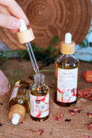 A hand holds a dropper over an open bottle of "Vanilla" tantric massage oil on a tree stump. Two other bottles are standing or lying next to it. Dried flower petals are scattered around the bottles.