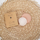 Eine rosa Menstruationsscheibe in Größe M, aus 100% medizinischem Silikon, wird zusammen mit ihrer braunen Kartonverpackung auf einer gewebten Matte präsentiert.