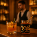 Ein Glas ANUK Manuka Honig MGO 400 mit einem schwarz-goldenen Etikett und schwarzem Deckel, das auf einem braunen Tuch neben einem kleinen goldenen Löffel liegt.