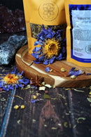 Zwei Teepackungen mit der Aufschrift "Third Eye Chakra Tea" stehen auf einem Holzbrett. Sie sind von blauen, getrockneten Lotusblumen, Sternanis und verstreuten Kräutern umgeben.