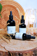 Drei schwarze Tropfflaschen mit Lion's Mane Flüssigextrakt auf einer Holzscheibe, eine liegend, mit getrockneten Gräsern und einer goldenen Drahtlampe.
