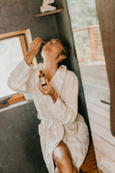 Eine Frau mit grauem Haar, bekleidet mit einem gestreiften weißen Bademantel, lehnt in einem Badezimmer an einer dunklen gefliesten Wand und tropft sich mit einer Pipette PAAVANI Nasenöl in die Nase, wobei sie nach oben blickt.