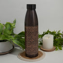 Verzierte dunkle Kupfer-Wasserflasche auf einem gewebten Untersetzer, neben einer brennenden Kerze und grünen Zimmerpflanzen.