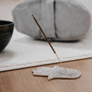 Ein angezündetes, braunes Räucherstäbchen, das sanft raucht, steckt in einem detaillierten silberfarbenen Hamsa-Hand-Räucherstäbchenhalter auf einem Holzboden. Im unscharfen Hintergrund sind eine dunkle Klangschale und ein hellgraues Meditationskissen zu sehen.