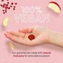 Eine Nahaufnahme, die betont, dass das Produkt 100% Vegan ist. Eine Hand mit lackierten Fingernägeln hält ein einzelnes quadratisches rotes Gummibärchen. Der Text besagt, dass die Gummibärchen mit natürlichem Fruchtsaft hergestellt werden. Apfelscheiben und Granatapfelkerne sind am Rand verstreut.