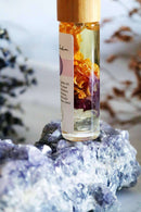 Eine Roll-on-Flasche mit Chakra-Öl und einem Holzdeckel steht auf einem leuchtend blauen und lila Fluoritkristall, der sich auf einer hellen Oberfläche befindet.