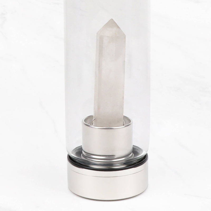 Eine Nahaufnahme des unteren Teils einer durchsichtigen Glaswasserflasche, die den Edelstahlsockel und einen prominenten, klaren Quarzkristall zeigt, der von diesem nach oben ragt. Die Flasche steht auf einer weißen Marmoroberfläche.