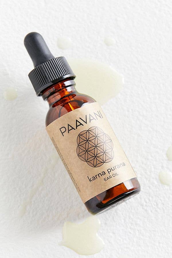 Eine schräg gestellte Flasche PAAVANI Karna Purana Ohrenöl auf einer weißen Oberfläche, umgeben von kleinen Ölpfützen, die die Textur des Produkts zeigen.