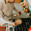 Aufnahme aus der Vogelperspektive eines Mannes, der einen Löffel Kurkumapulver aus einem Glas in einen Topf mit Milch auf dem Herd gibt, um Goldene Milch zuzubereiten.