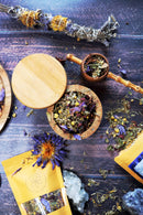 Eine Vogelperspektive zeigt eine offene Holzdose mit getrockneten Kräutern und Blüten. Um die Dose herum sind eine Teepackung, getrocknete Blumen und Kristalle auf einer rustikalen Holzoberfläche verstreut.