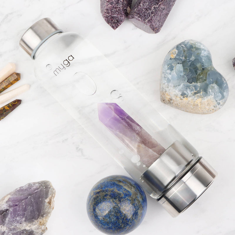 Eine transparente "myga" Wasserflasche mit einem Amethystkristall, umgeben von verschiedenen natürlichen Mineralien und Kristallen auf einer weißen Marmoroberfläche.