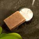 Ein Stück gesprenkelte braune Seife liegt auf einer nassen, dunklen Oberfläche in der Nähe eines Metallabflusses.