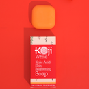 Orangefarbene Seife und rote Schachtel mit „Koji White Kojic Acid Skin Brightening Soap“ auf rotem Hintergrund. Die Schachtel zeigt Pflanzenillustrationen, die einen frischen, lebendigen Ton vermitteln.