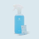 Eine Produktaufnahme auf hellblauem Hintergrund, die eine durchscheinende, mattierte Sprühflasche für COSMEAU GLAS-REINIGER mit einer leuchtend blauen Flüssigkeit zeigt. Vor der Flasche liegt ein einzelner versiegelter Folienbeutel mit GLAS BLAU Reinigungs-Tabs und einer passenden blauen, sprudelnden Tablette im Vordergrund.