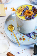 Eine Nahaufnahme einer goldenen Latte, die mit lila und orangefarbenen Trockenblumen auf einer blauen Untertasse garniert ist.