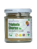 Ein Glasgefäß mit "Ayurveda autentico Triphala Churna Bio" (einer Mischung aus drei Früchten) und einem weißen Deckel. Das Etikett zeigt grüne Schrift und Symbole für "Sin Gluten" (glutenfrei), "100% Natur", "OMG" (gentechnikfrei) und "150 g".