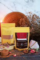 Eine dunkelrote und gelbe Packung „Love Choc Aphrodisiac & Adaptogenic Cacao Elixir“ steht im Vordergrund neben einem passenden gelben Beutel, umgeben von Kakaopulver, Rosenblättern, einem rosa Rosenquarzkristall und einer brennenden Kerze.