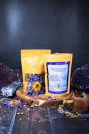 Zwei Teepackungen, von denen eine als "Third Eye Chakra Tea" gekennzeichnet ist, stehen auf einer Holzplatte. Sie sind umgeben von blauen und gelben getrockneten Blumen und Kristallen vor einem dunklen Hintergrund.