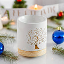 Eine weiße Keramik-Duftlampe oder Aromalampe mit einem ausgeschnittenen Lebensbaum-Motiv, umgeben von blauen Weihnachtskugeln, Tannenzweigen und einer brennenden Kerze.