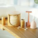 Eine Sammlung von Bamboozy Nachhaltigkeitsprodukten zur Körperpflege auf einer Holzfläche, darunter ein Wattepadhalter, zwei Wattestäbchenbehälter, ein roségoldener Rasierhobel und eine rosa Menstruationstasse.