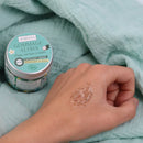 Eine Hand mit einer Probe des Aumyana Gommage Aloha Gesichtspeelings neben dem Tiegel.