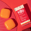 Orangefarbene Seife und rote Schachtel mit „Koji White Kojic Acid Skin Brightening Soap“ auf rotem Hintergrund.