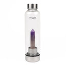 Eine transparente "myga" Wasserflasche mit einem spitz zulaufenden violetten Amethystkristall auf einem silbernen Sockel, stehend auf einer weißen Marmoroberfläche.