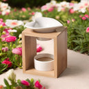 Eine Aromalampe aus Bambusholz mit einer herzförmigen Keramikschale, draußen inmitten von rosa und weißen Blumen.