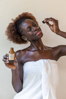 Eine dunkelhäutige Frau mit lockigem braunem Haar, eingewickelt in ein weißes Handtuch, hält in ihrer linken Hand eine Flasche "PAAVANI AYURVEDA NOSE OIL" und trägt mit einer Pipette in ihrer rechten Hand Öl auf ihre Nase auf, die Augen konzentriert geschlossen.