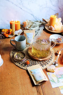 Ein Teetisch mit einer Glaskanne, die Kräutertee enthält, umgeben von Kerzen, getrockneten Blumen, einer Tasse, zwei Teepackungen und Tarotkarten.