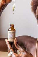 Eine Person hält eine Flasche Paavani Ayurveda Tridoshic Serum, während ein Tropfen Öl aus der erhobenen Pipette fällt.