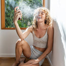 Eine lächelnde Frau mit lockigem, blondem Haar sprüht sich das Paavani Rosenwasser auf ihr Gesicht, während sie im Sonnenlicht auf dem Boden sitzt, mit einem Fenster und Pflanzen im Hintergrund.