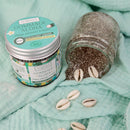 Ein Tiegel Aumyana Gommage Aloha Gesichtspeeling neben einem umgekippten Glas mit Peeling und Kaurischnecken.