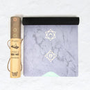 Eine teilweise ausgerollte Myga Yogamatte aus veganem Wildleder auf Gummi mit einem hellvioletten Marmordesign und zwei goldfarbenen Chakra- oder Himmelssymbolen, die neben ihrer Verpackungsröhre liegt.