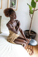 Eine dunkelhäutige Frau mit lockigem braunem Haar in einem goldenen Kleid sitzt auf einem weißen Bett und trägt Körperöl auf ihren rechten Arm auf. Eine Flasche PAAVANI-Öl steht auf einem dunklen Hocker neben ihr.