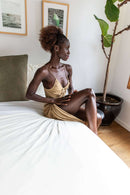  Eine dunkelhäutige Frau mit lockigem braunem Haar in einem goldenen Spaghettiträgerkleid sitzt seitlich auf einem weißen Bett und blickt nach rechts. Ihre rechte Hand ruht sanft auf ihrem linken Knie.