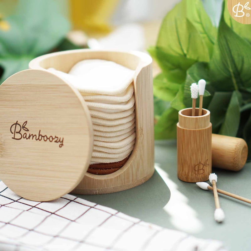 Wiederverwendbare Wattestäbchen von Bamboozy in einem Bambushalter, zusammen mit einem gemusterten Tuch und grünen Blättern, alles auf einer grauen Oberfläche.
