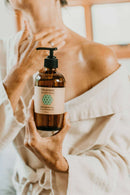 Eine Frau in einem weißen Bademantel hält eine Flasche PAAVANI Vata Körperöl mit einem grünen geometrischen Muster in einer Hand, während sie mit der anderen sanft ihren Nacken und ihre Schulter massiert, vermutlich trägt sie das Öl auf.